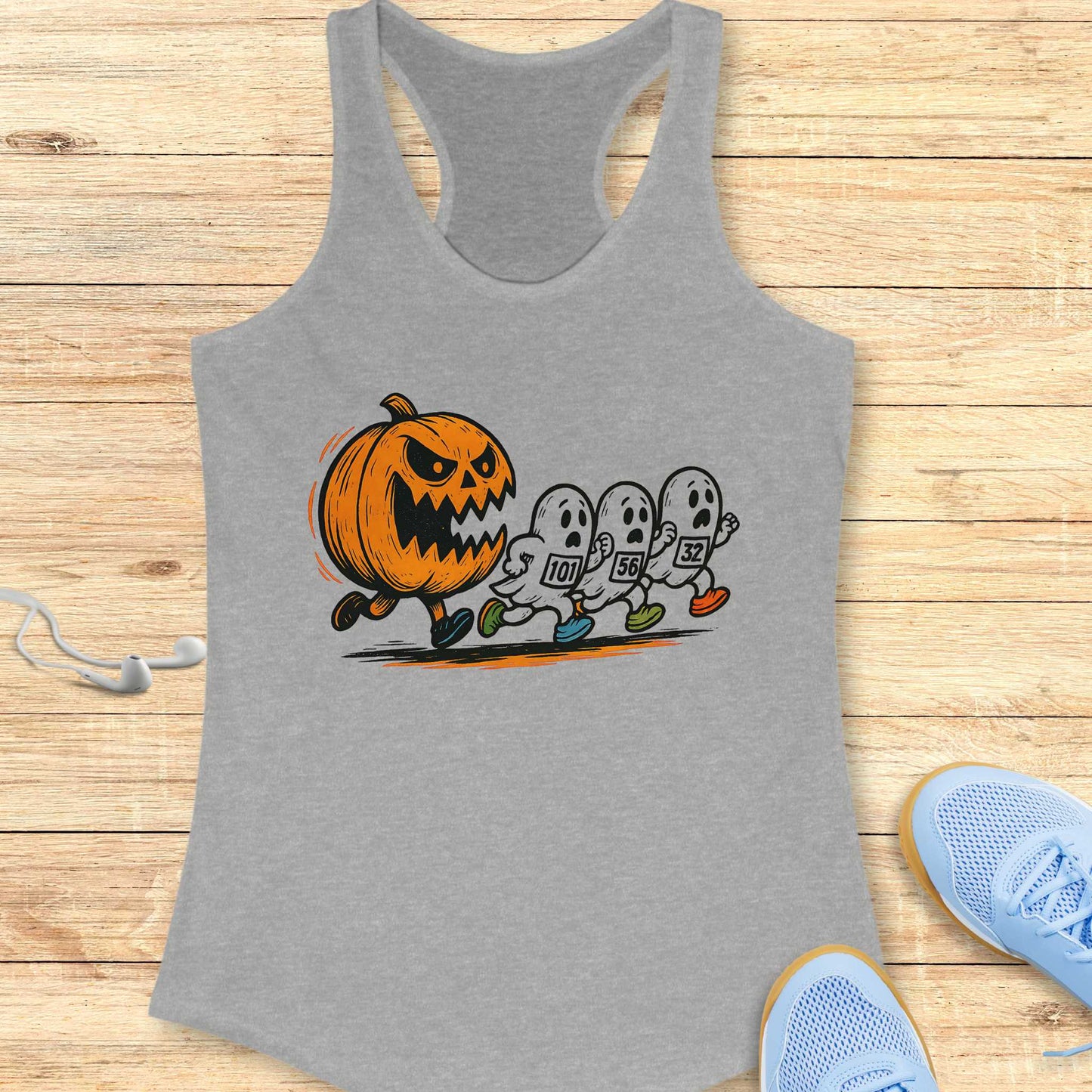 Grey racerback tank top featuring a vibrant illustration of a menacing orange pumpkin chasing three white running ghosts wearing colorful shoes and race bibs, displayed on a wooden surface beside white earbuds and light blue athletic sneakers