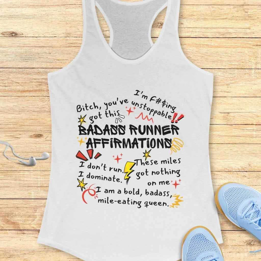 White racerback tank top displaying bold, colorful "BADASS RUNNER AFFIRMATIONS" text with doodle-style graphics, laid on a light wooden background with light blue running shoes and white earphones visible