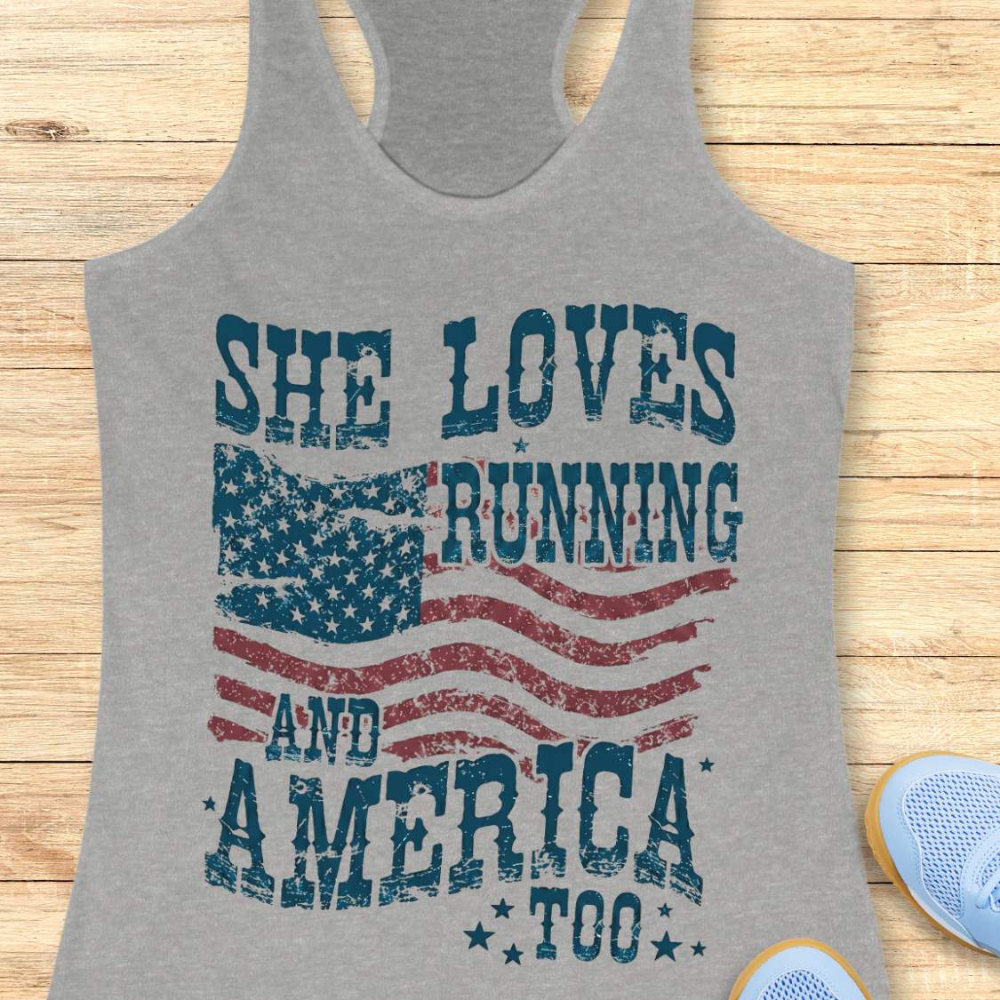 Gray tank top with 'She Loves Running and America Too' text and American flag design on a wooden surface.
