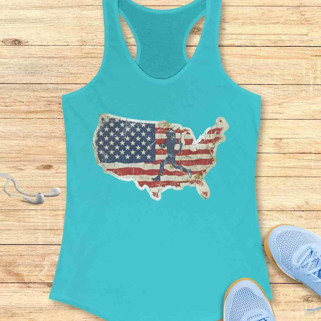 A turquoise racerback tank top features a distressed American flag map with a running figure silhouette, displayed on a wooden surface with light blue athletic shoes