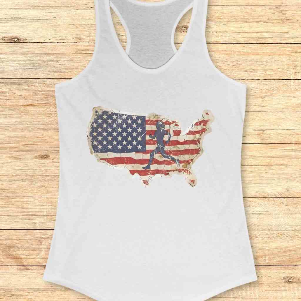 White racerback tank top with a distressed United States map graphic filled with a weathered American flag pattern and a dark silhouette of a runner, presented on a rustic wooden plank surface