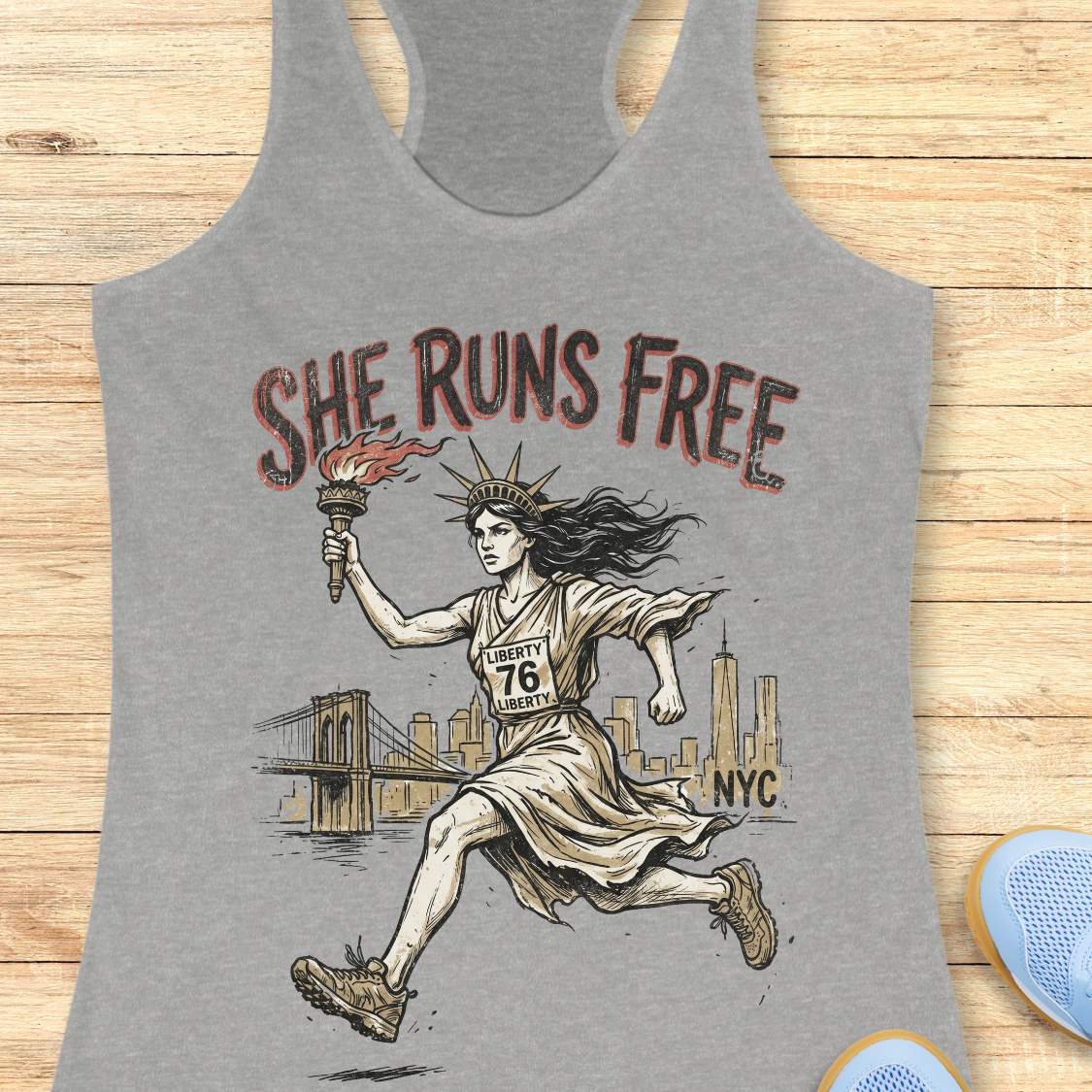 grey tank top with a design of lady liberty running and text - 'she runs free NYC', on a wooden surface.