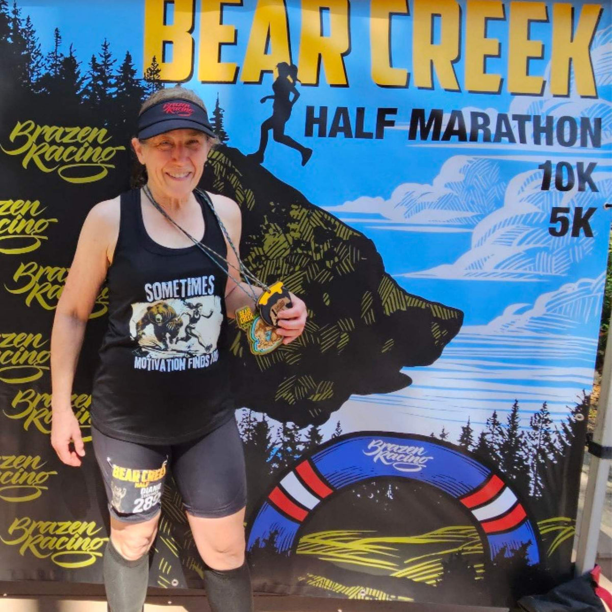 A woman stands in front of a Bear Creek Half Marathon 10K and 5K sign, wearing a black tank top with a 'Sometimes Motivation Forgot' graphic, black shorts, and a black cap. She holds a race medal.