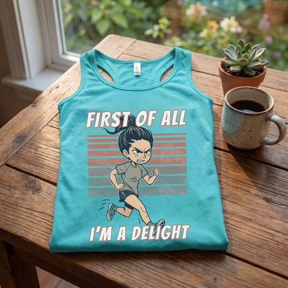 A turquoise tank top laid flat on a wooden table, featuring a cartoon running man graphic with the text 'FIRST OF ALL I'M A DELIGHT'. A ceramic mug of coffee and a small potted succulent sit beside it, with a window and garden visible in the background.