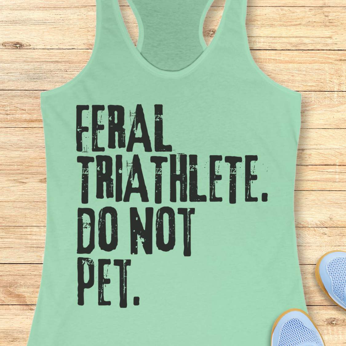 mint racerback tank top displayed on a light wood background, featuring bold distressed white lettering that reads: “FERAL TRIATHLETE. DO NOT PET.” The minimalist design uses large vintage-style typography for a humorous triathlon-themed statement. Partial blue running shoes are visible in the bottom corners of the image.