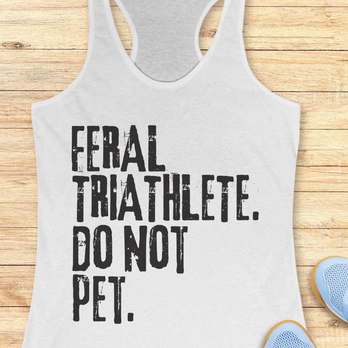 white racerback tank top displayed on a light wood background, featuring bold distressed white lettering that reads: “FERAL TRIATHLETE. DO NOT PET.” The minimalist design uses large vintage-style typography for a humorous triathlon-themed statement. Partial blue running shoes are visible in the bottom corners of the image.