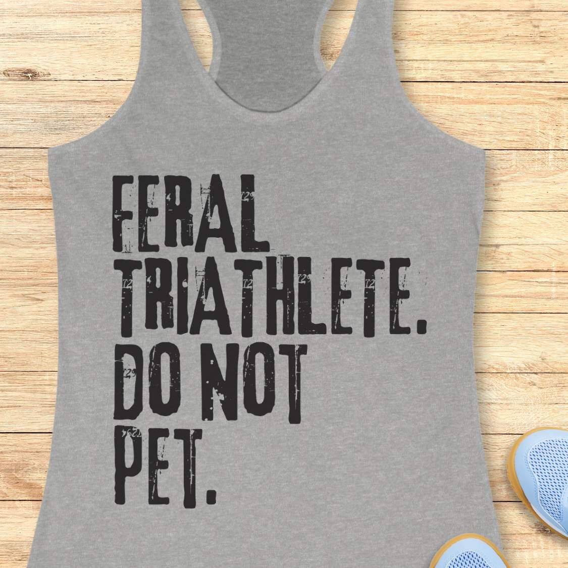 grey racerback tank top displayed on a light wood background, featuring bold distressed white lettering that reads: “FERAL TRIATHLETE. DO NOT PET.” The minimalist design uses large vintage-style typography for a humorous triathlon-themed statement. Partial blue running shoes are visible in the bottom corners of the image.