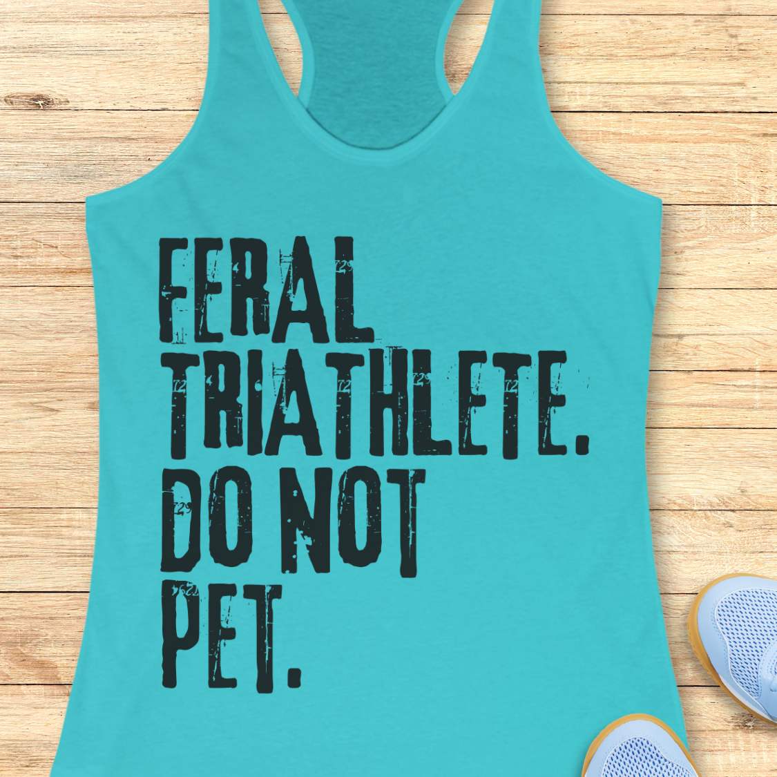 blue racerback tank top displayed on a light wood background, featuring bold distressed white lettering that reads: “FERAL TRIATHLETE. DO NOT PET.” The minimalist design uses large vintage-style typography for a humorous triathlon-themed statement. Partial blue running shoes are visible in the bottom corners of the image.