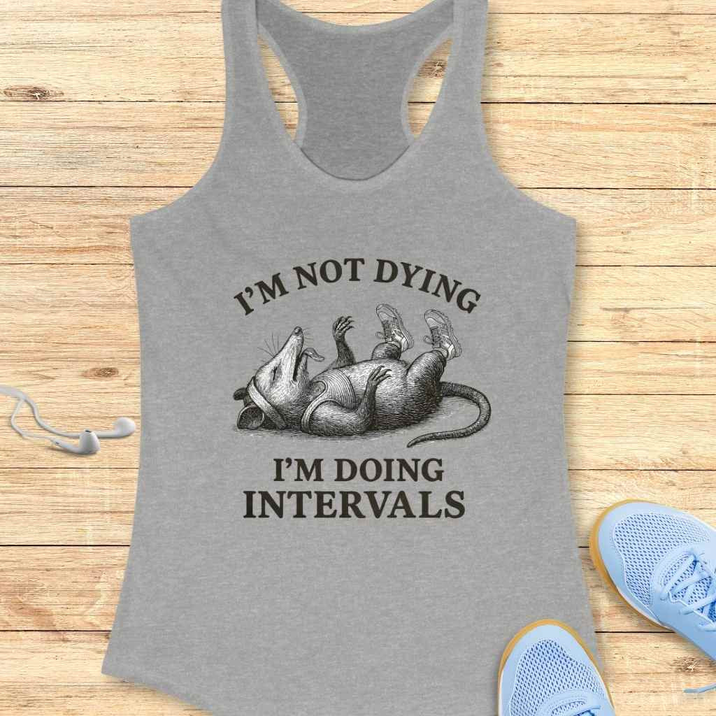 Grey racerback tank top featuring a black and white graphic of an opossum wearing sneakers and a headband, lying on its back with the text I'M NOT DYING I'M DOING INTERVALS, displayed on a rustic wooden background with white earbuds and blue athletic shoes partially visible