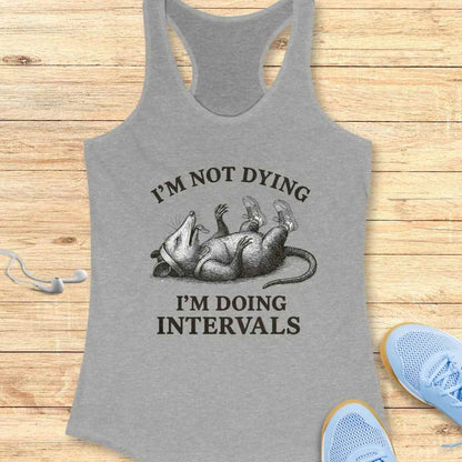 Grey racerback tank top featuring a black and white graphic of an opossum wearing sneakers and a headband, lying on its back with the text I'M NOT DYING I'M DOING INTERVALS, displayed on a rustic wooden background with white earbuds and blue athletic shoes partially visible