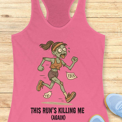pink racerback tank top displaying a humorous cartoon zombie runner with green skin, tattered brown clothes, race bibs 131 and 1313, and the slogan 'THIS RUN'S KILLING ME (AGAIN)' set against a light wooden background with partial blue running shoes and white earbuds visible