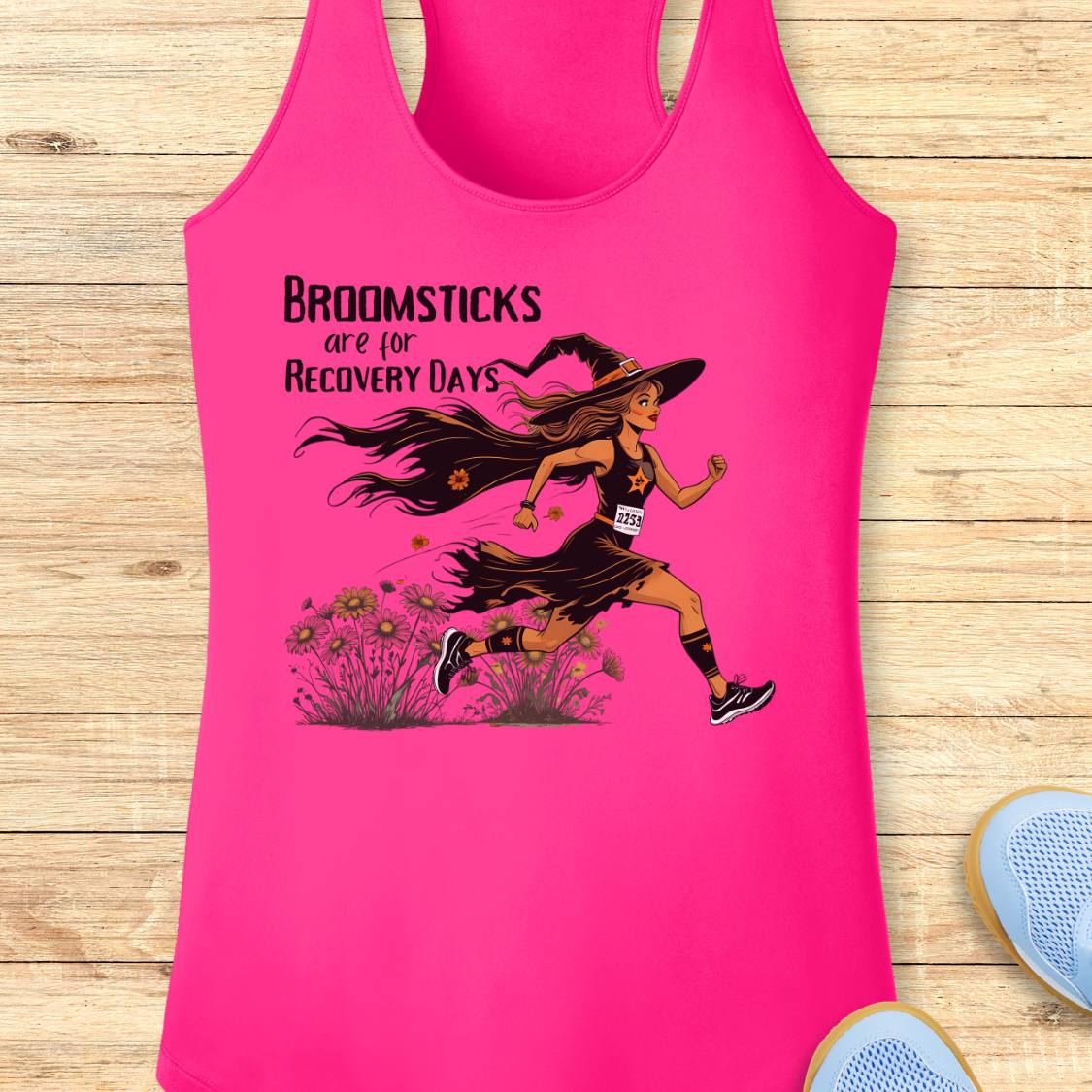 A bright pink racerback tank top laid flat on a light wood plank background. The tank top features a graphic of a witch running with broomsticks and the text 'BROOMSTICKS are for RECOVERY DAYS'.