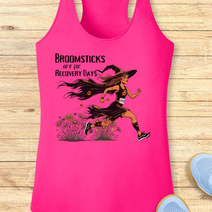 A bright pink racerback tank top laid flat on a light wood plank background. The tank top features a graphic of a witch running with broomsticks and the text 'BROOMSTICKS are for RECOVERY DAYS'.