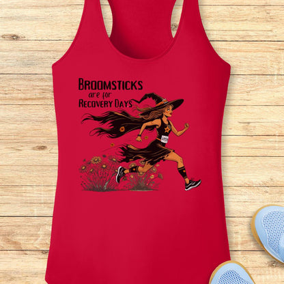 A red racerback tank top laid flat on a light wood background, featuring a graphic of a witch running with the text 'BROOMSTICKS are for RECOVERY DAYS.' Part of a blue shoe is visible at the bottom right as a styling prop.