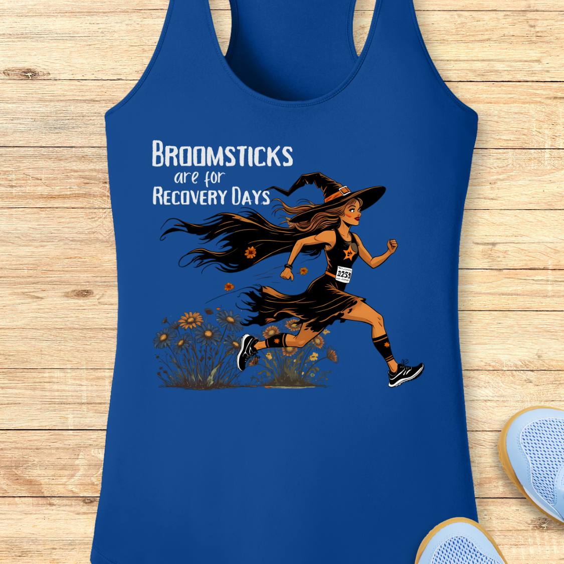 A royal blue racerback tank top laid flat on a light wood background, featuring a graphic of a witch running with the text 'BROOMSTICKS are for RECOVERY DAYS.' A partial white shoe is visible at the bottom right as a styling prop.