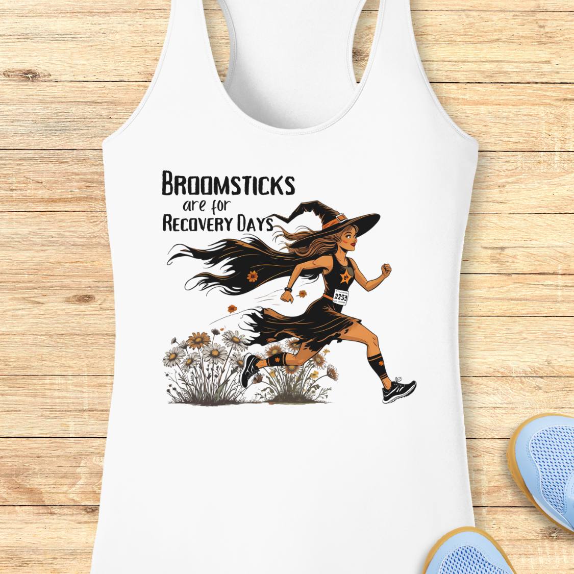 A white racerback tank top laid flat on a light wood surface, featuring a graphic of a woman running with broomsticks and the text 'Broomsticks are for Recovery Day.' Part of a blue shoe is visible at the bottom right.