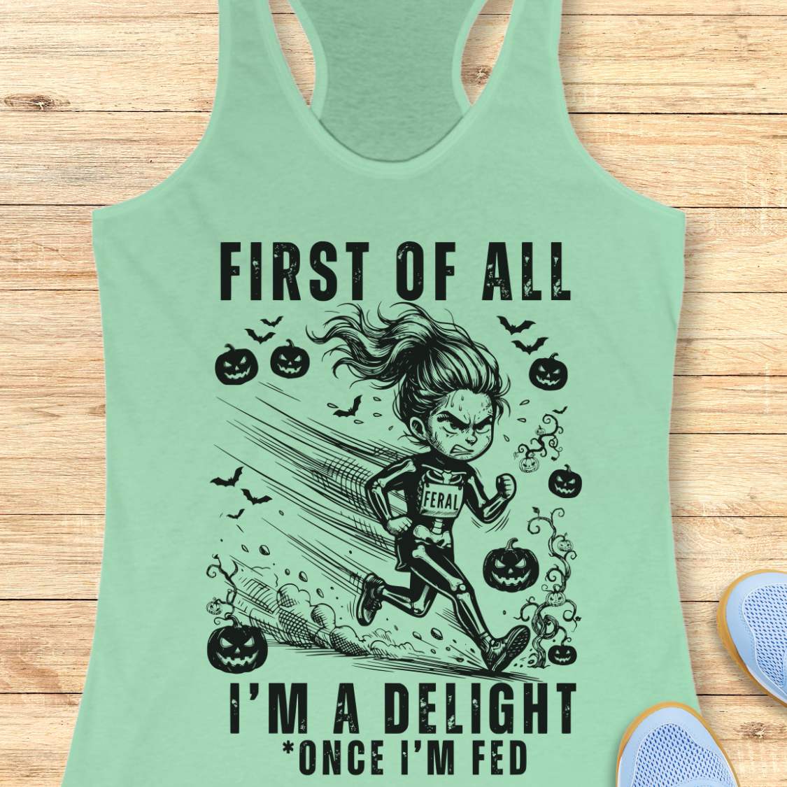 A mint green racerback tank top with a black graphic of a girl running and the text 'FIRST OF ALL I'M A DELIGHT * ONCE I'M FED', displayed flat on a light wood background with partial shoes visible at the edges.