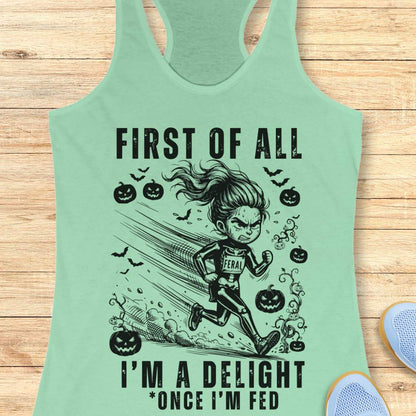 A mint green racerback tank top with a black graphic of a girl running and the text 'FIRST OF ALL I'M A DELIGHT * ONCE I'M FED', displayed flat on a light wood background with partial shoes visible at the edges.