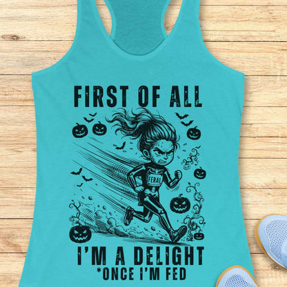 A turquoise racerback tank top laid flat on a light wood surface, featuring a black graphic of a woman running with pumpkins, bats, and the text 'FIRST OF ALL I'M A DELIGHT *ONCE I'M FED'. Part of a white shoe is visible at the bottom right.