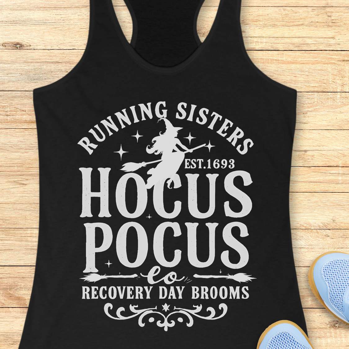 black tank top with witch graphic design and text 'running sisters est. 1693 hocus pocus recovery day brooms.', on a wooden surface