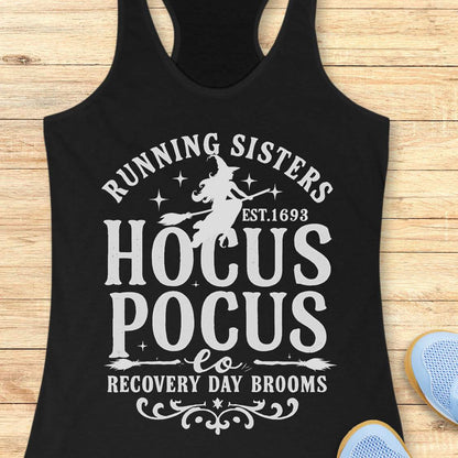 black tank top with witch graphic design and text 'running sisters est. 1693 hocus pocus recovery day brooms.', on a wooden surface