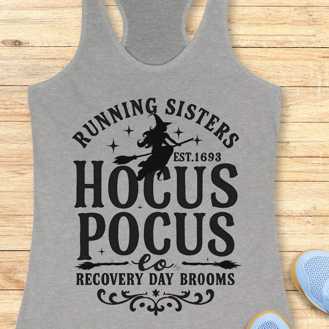 grey tank top with witch graphic design and text 'running sisters est. 1693 hocus pocus recovery day brooms.', on a wooden surface