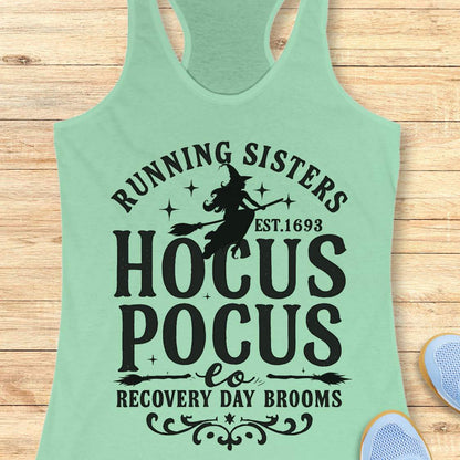 mint tank top with witch graphic design and text 'running sisters est. 1693 hocus pocus recovery day brooms.', on a wooden surface