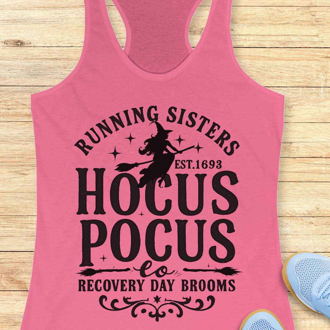pink tank top with witch graphic design and text 'running sisters est. 1693 hocus pocus recovery day brooms.', on a wooden surface