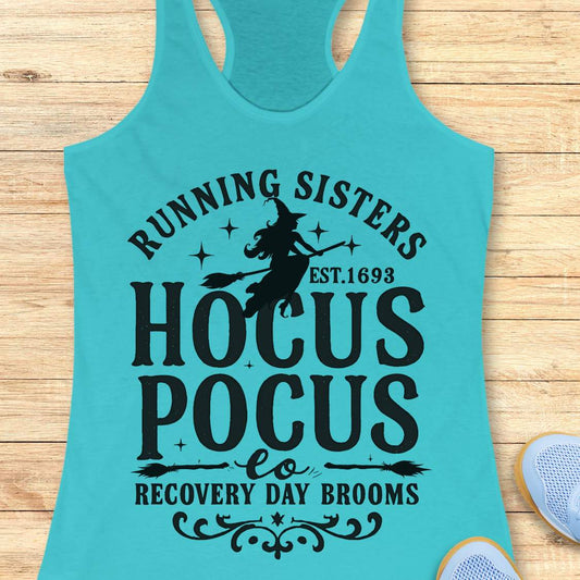 blue tank top with witch graphic design and text 'running sisters est. 1693 hocus pocus recovery day brooms.', on a wooden surface