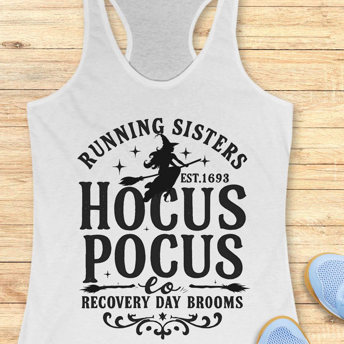 white tank top with witch graphic design and text 'running sisters est. 1693 hocus pocus recovery day brooms.', on a wooden surface