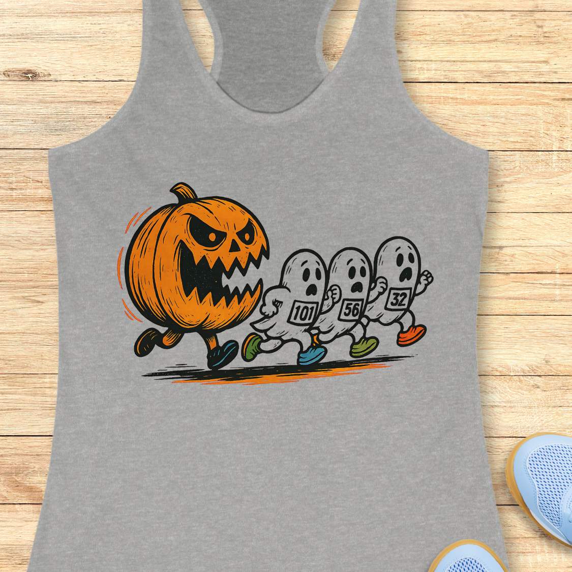 grey racerback tank top featuring a Halloween running graphic of an angry orange jack-o'-lantern chasing three white ghosts wearing numbered race bibs 101 56 and 32 with colorful blue green and orange running shoes displayed on a light wooden background alongside white earbuds and light blue sneakers