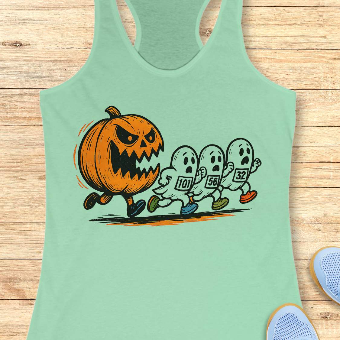 mint racerback tank top featuring a Halloween running graphic of an angry orange jack-o'-lantern chasing three white ghosts wearing numbered race bibs 101 56 and 32 with colorful blue green and orange running shoes displayed on a light wooden background alongside white earbuds and light blue sneakers