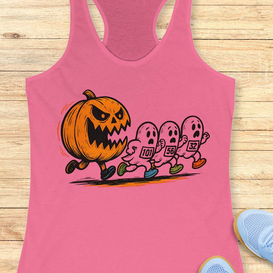 pink racerback tank top featuring a Halloween running graphic of an angry orange jack-o'-lantern chasing three white ghosts wearing numbered race bibs 101 56 and 32 with colorful blue green and orange running shoes displayed on a light wooden background alongside white earbuds and light blue sneakers