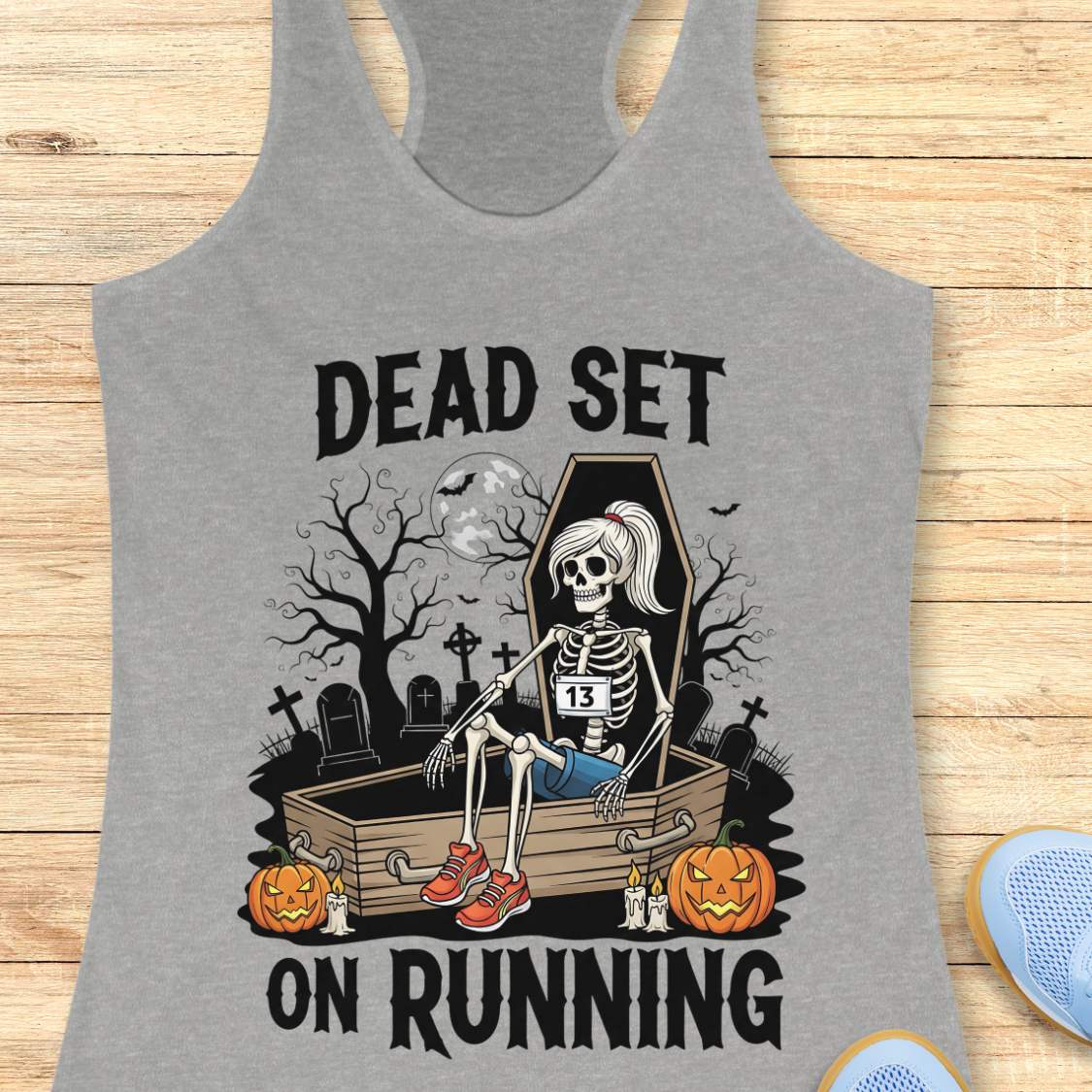 Illustration on a grey tank top depicting a female skeleton runner with a ponytail, a number 13 bib, blue shorts, and red sneakers, sitting in an open coffin in a spooky graveyard with bare trees, tombstones, and a full moon, flanked by two glowing jack-o'-lanterns, with the text Dead Set On Running
