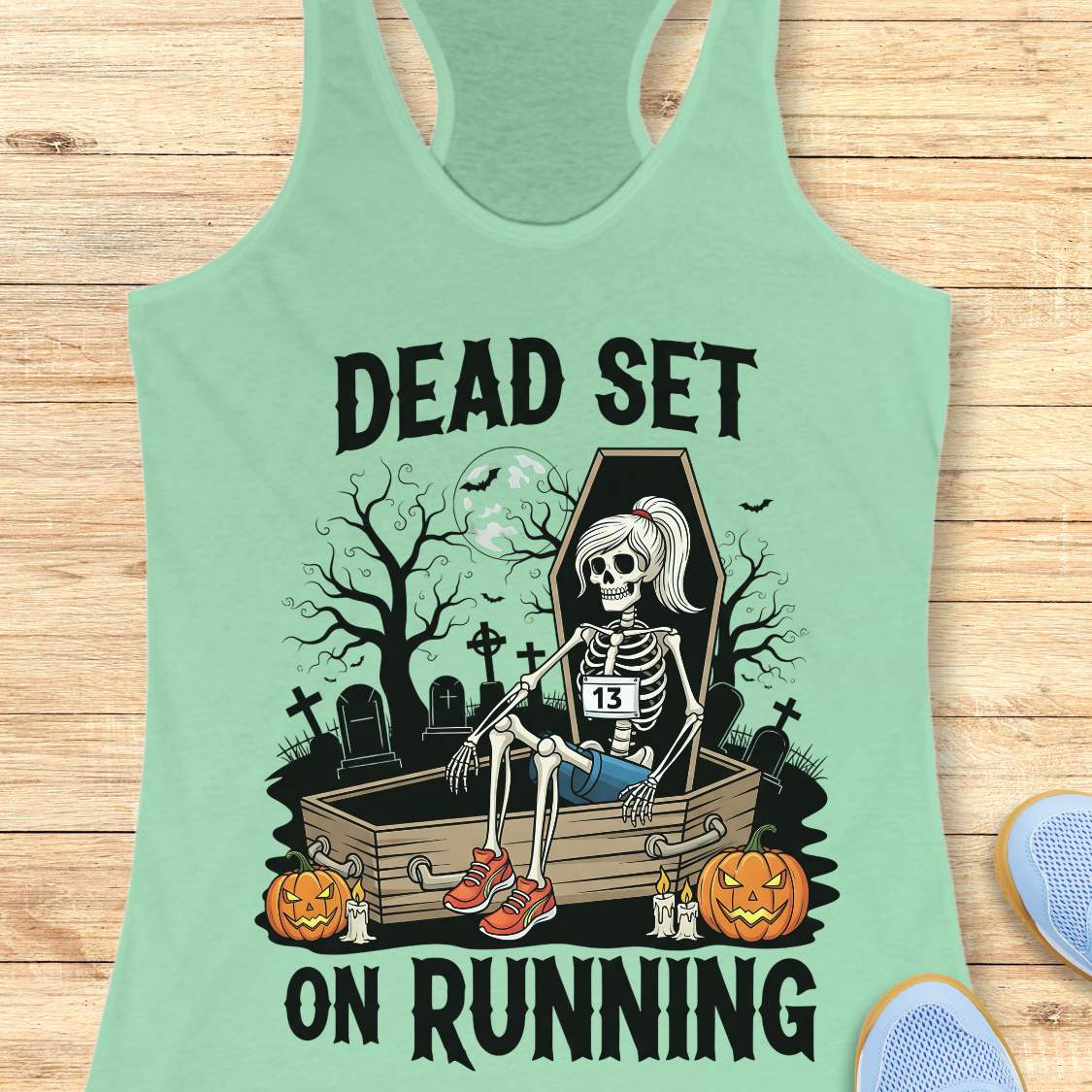 Illustration on a mint tank top depicting a female skeleton runner with a ponytail, a number 13 bib, blue shorts, and red sneakers, sitting in an open coffin in a spooky graveyard with bare trees, tombstones, and a full moon, flanked by two glowing jack-o'-lanterns, with the text Dead Set On Running