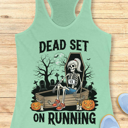 Illustration on a mint tank top depicting a female skeleton runner with a ponytail, a number 13 bib, blue shorts, and red sneakers, sitting in an open coffin in a spooky graveyard with bare trees, tombstones, and a full moon, flanked by two glowing jack-o'-lanterns, with the text Dead Set On Running