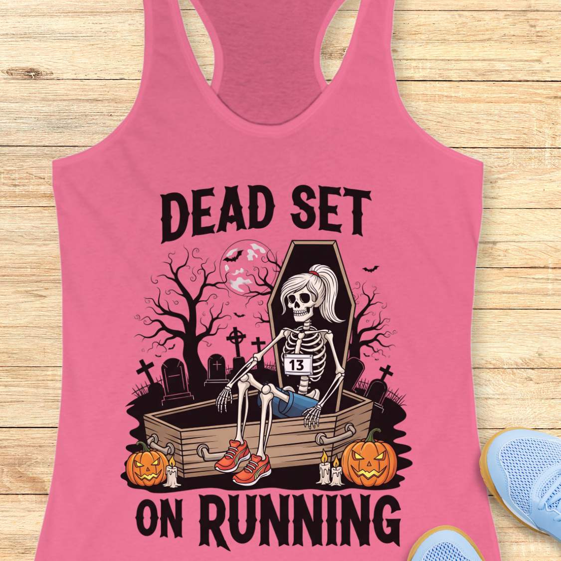 Illustration on a pink tank top depicting a female skeleton runner with a ponytail, a number 13 bib, blue shorts, and red sneakers, sitting in an open coffin in a spooky graveyard with bare trees, tombstones, and a full moon, flanked by two glowing jack-o'-lanterns, with the text Dead Set On Running