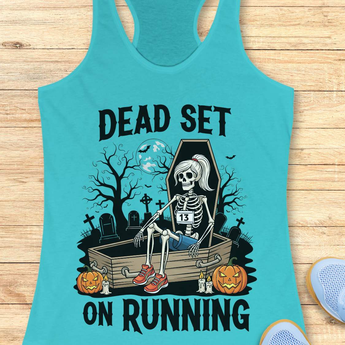 Illustration on a blue tank top depicting a female skeleton runner with a ponytail, a number 13 bib, blue shorts, and red sneakers, sitting in an open coffin in a spooky graveyard with bare trees, tombstones, and a full moon, flanked by two glowing jack-o'-lanterns, with the text Dead Set On Running