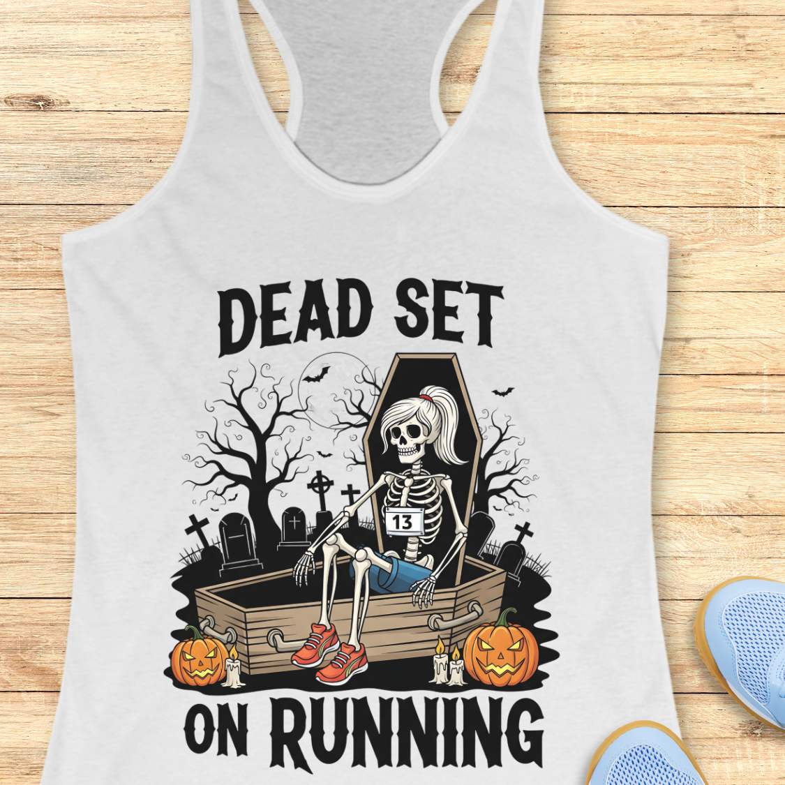 Illustration on a white tank top depicting a female skeleton runner with a ponytail, a number 13 bib, blue shorts, and red sneakers, sitting in an open coffin in a spooky graveyard with bare trees, tombstones, and a full moon, flanked by two glowing jack-o'-lanterns, with the text Dead Set On Running