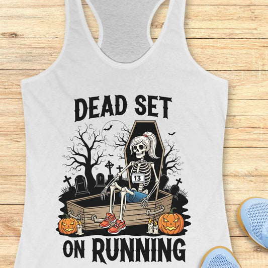 Illustration on a white tank top depicting a female skeleton runner with a ponytail, a number 13 bib, blue shorts, and red sneakers, sitting in an open coffin in a spooky graveyard with bare trees, tombstones, and a full moon, flanked by two glowing jack-o'-lanterns, with the text Dead Set On Running