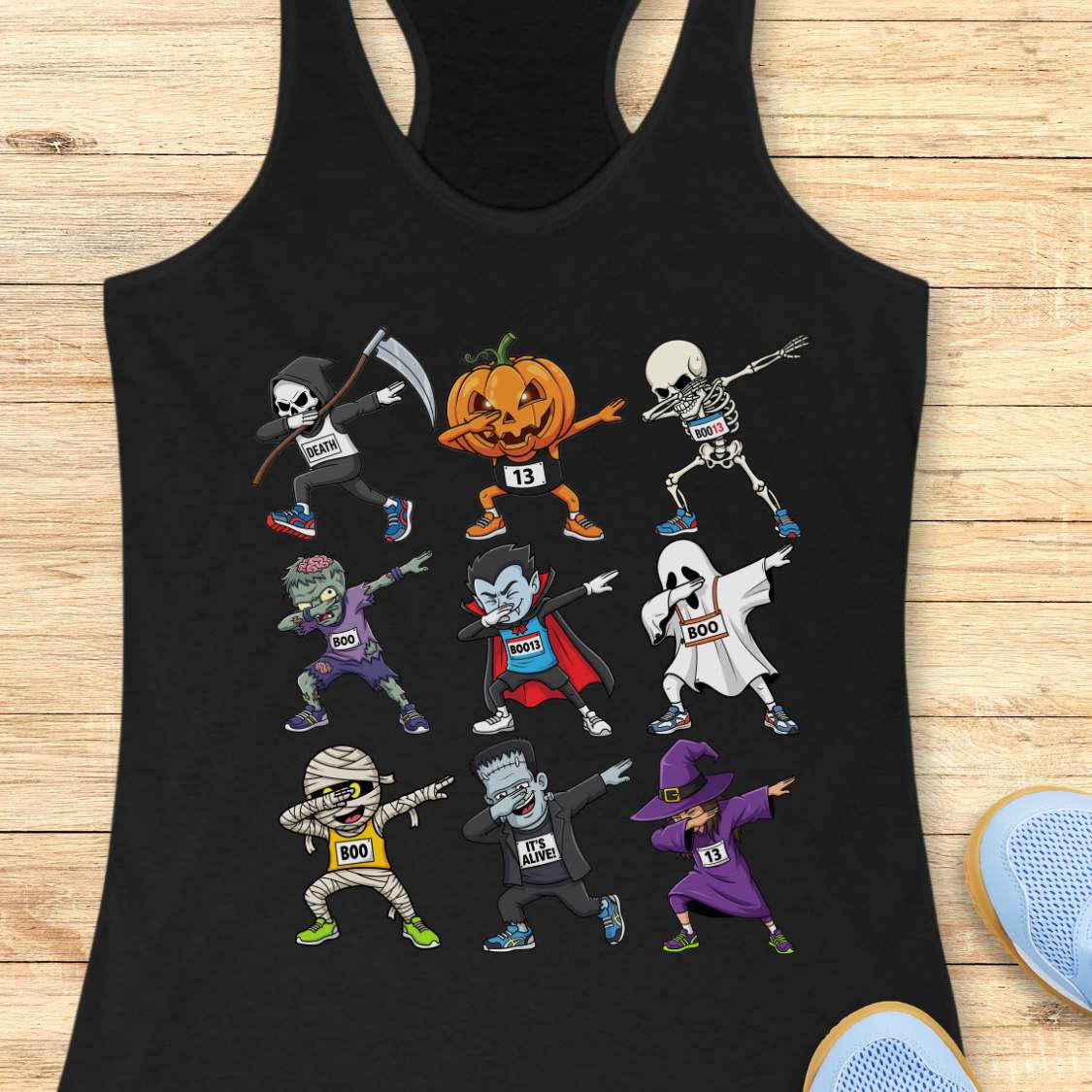 Digital illustration on a black racerback tank top showcasing nine dabbing Halloween cartoon characters including a Grim Reaper, pumpkin, skeleton, zombie, vampire, ghost, mummy, Frankenstein's monster, and a witch, many wearing race bibs like 'DEATH' or 'BOO,' displayed on a rustic wooden background with blue sneakers and white earbuds nearby