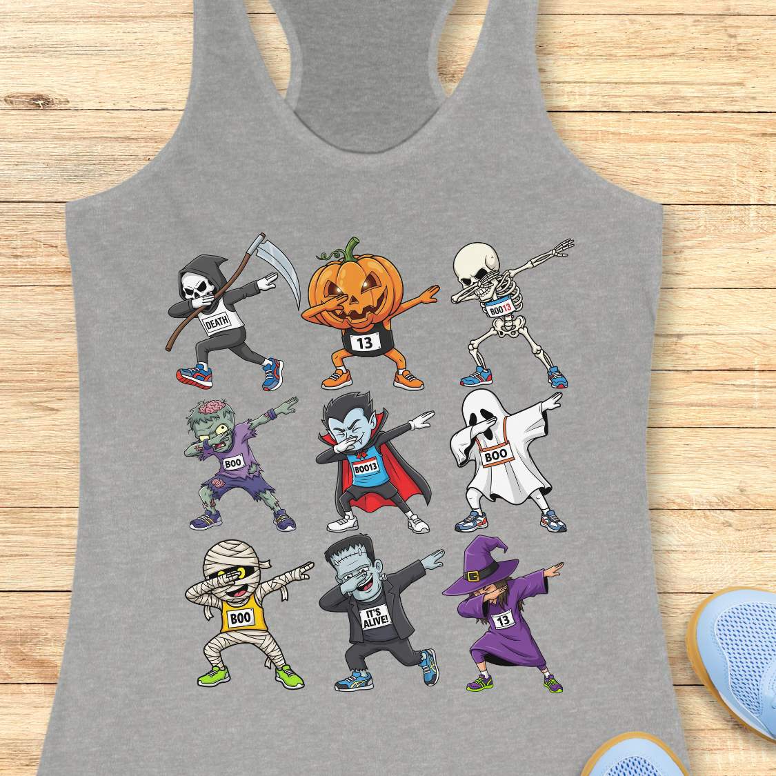 Digital illustration on a grey racerback tank top showcasing nine dabbing Halloween cartoon characters including a Grim Reaper, pumpkin, skeleton, zombie, vampire, ghost, mummy, Frankenstein's monster, and a witch, many wearing race bibs like 'DEATH' or 'BOO,' displayed on a rustic wooden background with blue sneakers and white earbuds nearby