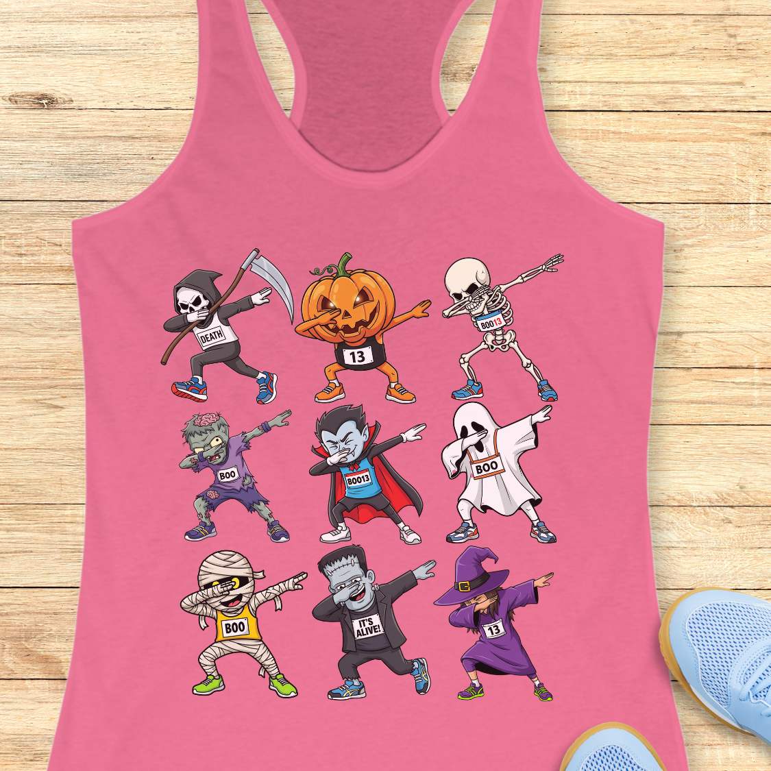 Digital illustration on a pink racerback tank top showcasing nine dabbing Halloween cartoon characters including a Grim Reaper, pumpkin, skeleton, zombie, vampire, ghost, mummy, Frankenstein's monster, and a witch, many wearing race bibs like 'DEATH' or 'BOO,' displayed on a rustic wooden background with blue sneakers and white earbuds nearby