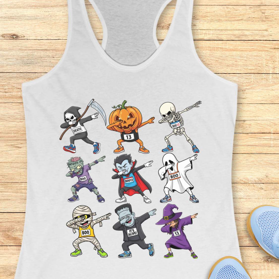 Digital illustration on a white racerback tank top showcasing nine dabbing Halloween cartoon characters including a Grim Reaper, pumpkin, skeleton, zombie, vampire, ghost, mummy, Frankenstein's monster, and a witch, many wearing race bibs like 'DEATH' or 'BOO,' displayed on a rustic wooden background with blue sneakers and white earbuds nearby