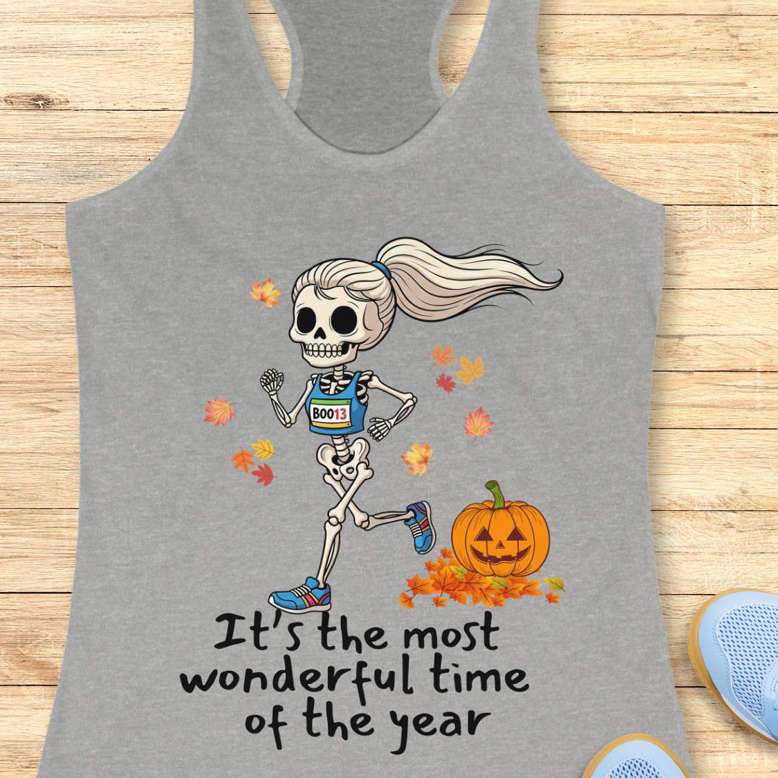 Graphic design on a grey tank top features a female skeleton with a ponytail wearing a blue running bib B0013 and blue sneakers, running past a smiling jack-o'-lantern and colorful autumn leaves, with the text It's the most wonderful time of the year