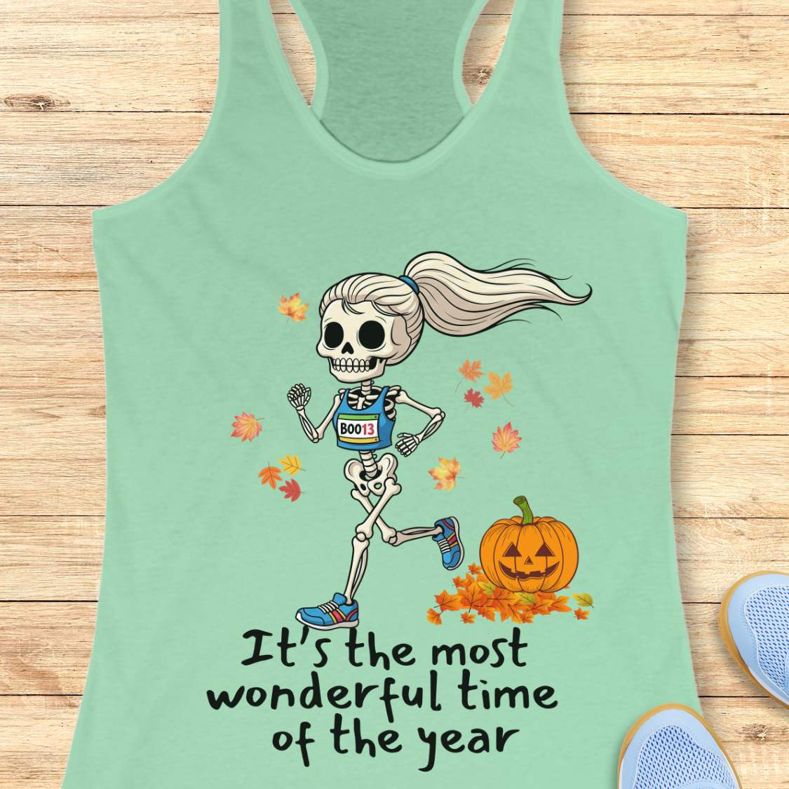 Graphic design on a mint tank top features a female skeleton with a ponytail wearing a blue running bib B0013 and blue sneakers, running past a smiling jack-o'-lantern and colorful autumn leaves, with the text It's the most wonderful time of the year