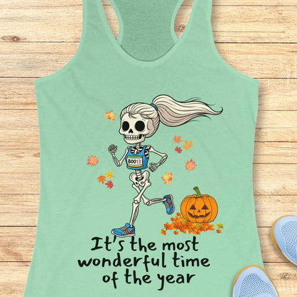 Graphic design on a mint tank top features a female skeleton with a ponytail wearing a blue running bib B0013 and blue sneakers, running past a smiling jack-o'-lantern and colorful autumn leaves, with the text It's the most wonderful time of the year