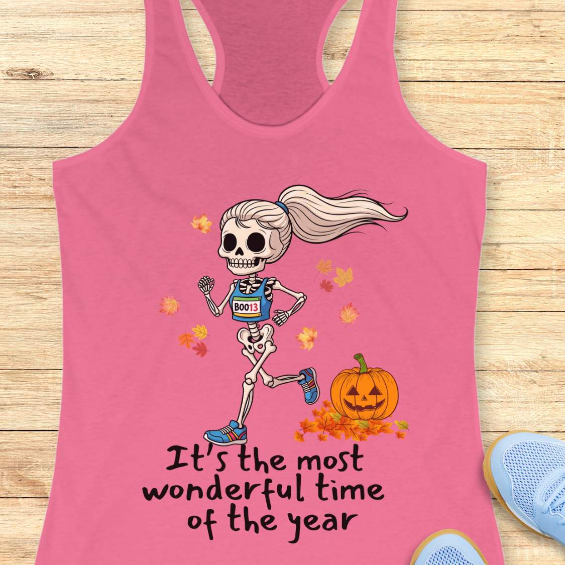 Graphic design on a pink tank top features a female skeleton with a ponytail wearing a blue running bib B0013 and blue sneakers, running past a smiling jack-o'-lantern and colorful autumn leaves, with the text It's the most wonderful time of the year