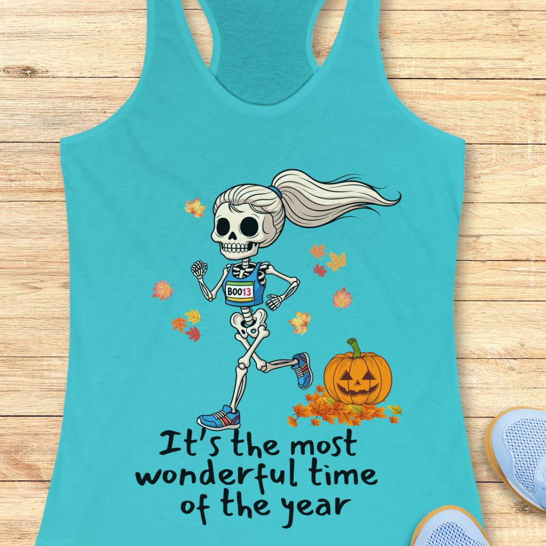 Graphic design on a blue tank top features a female skeleton with a ponytail wearing a blue running bib B0013 and blue sneakers, running past a smiling jack-o'-lantern and colorful autumn leaves, with the text It's the most wonderful time of the year