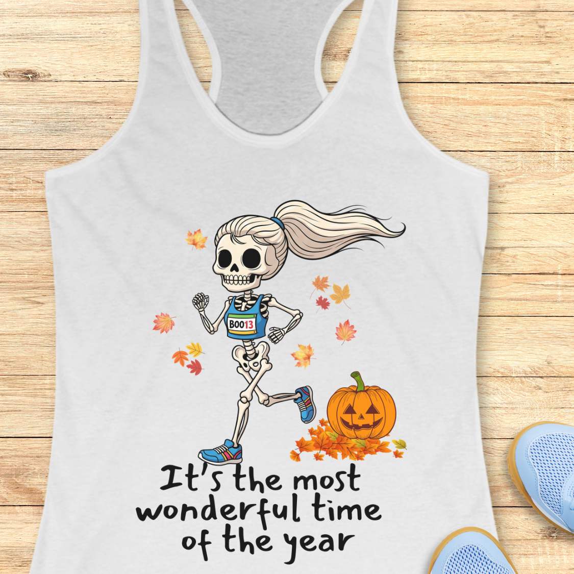 Graphic design on a white tank top features a female skeleton with a ponytail wearing a blue running bib B0013 and blue sneakers, running past a smiling jack-o'-lantern and colorful autumn leaves, with the text It's the most wonderful time of the year